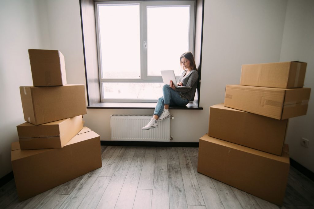 woman moving, sitting among cardboard boxes, using a laptop and smiling