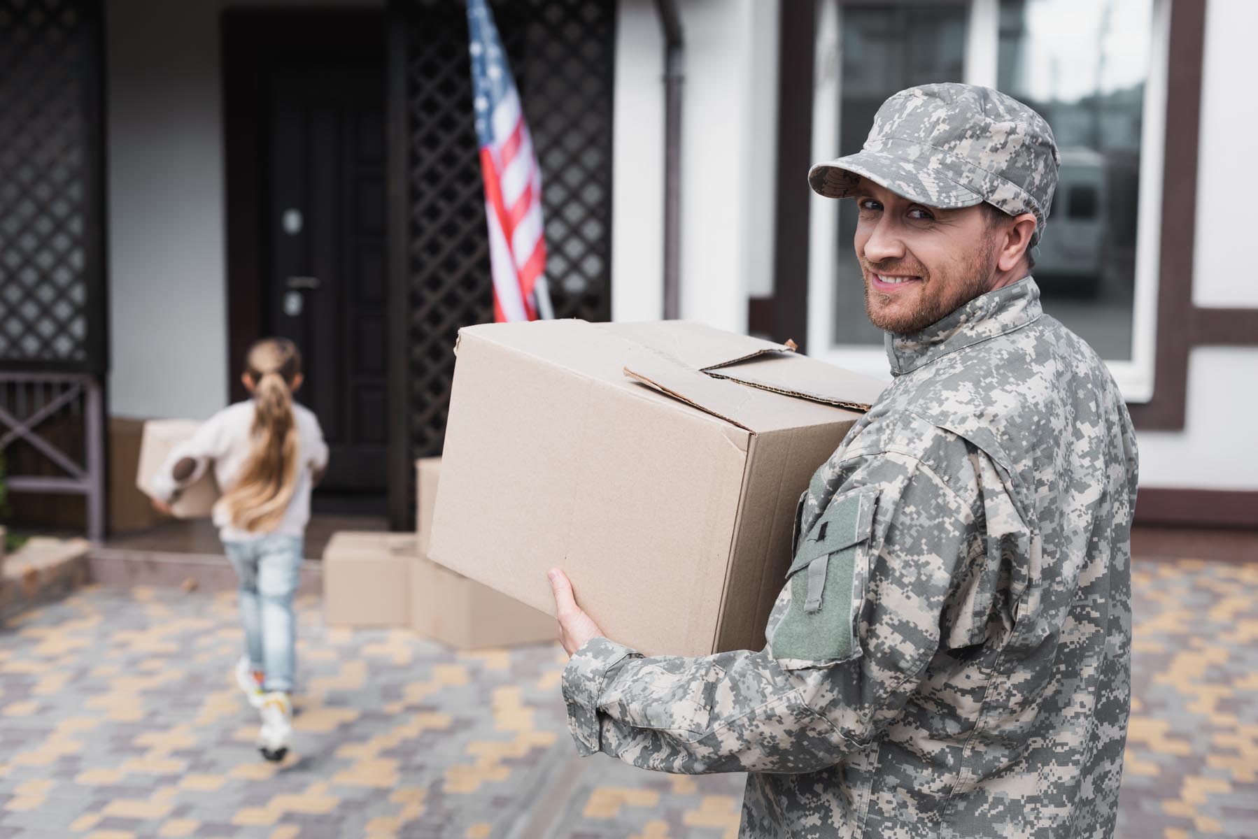 Happy military serviceman holding cardboard box and looking at camera with blurred girl on background