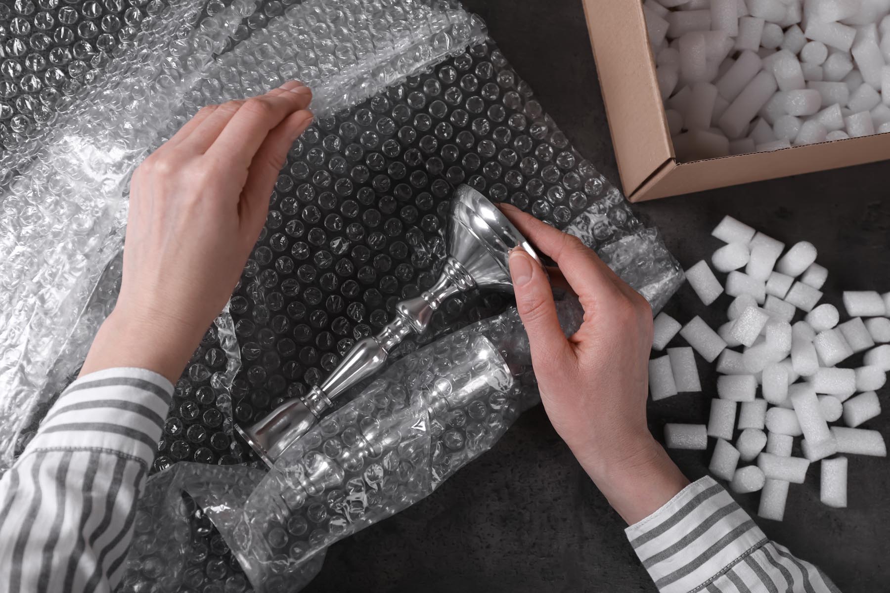 Woman covering silver candlesticks with bubble wrap at dark grey table, closeup