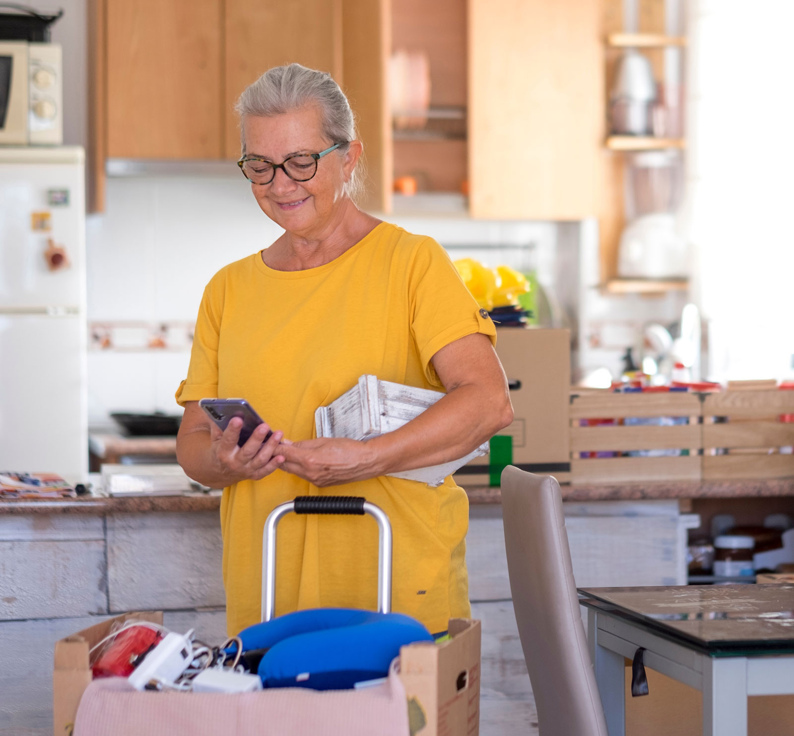 Senior woman looking at smart phone during preparation of relocation with a white small basket under the arm - lot of moving boxes and things in the kitchen